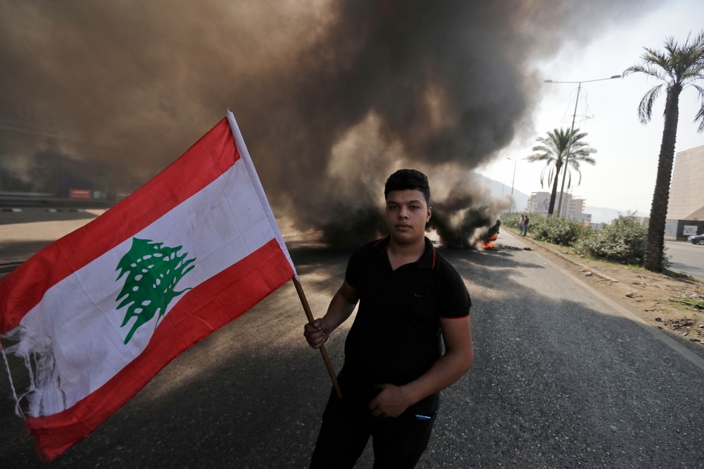 A Lebanese youth waves a national flag in front of burning tires blocking the Beirut-Tripoli highway in the coastal town of Ghazir, north of the port city of Junieh, during continuing anti-government protests across Lebanon on Wednesday. -AFP