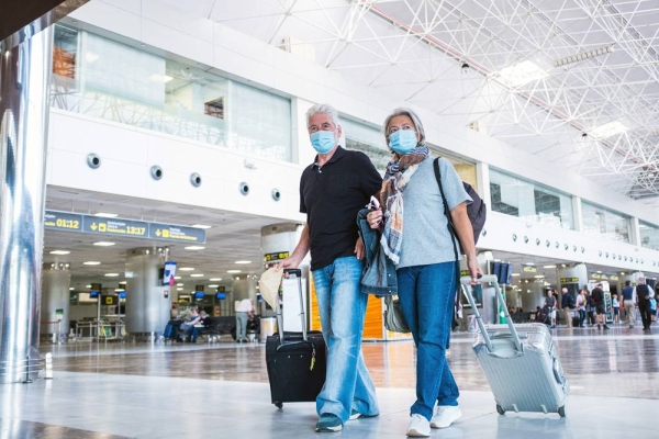 Couple of two seniors or mature people walking in the airport going to their gate and take their flight wearing medical mask to prevent virus like Coronavirus carrying luggage or trolley.