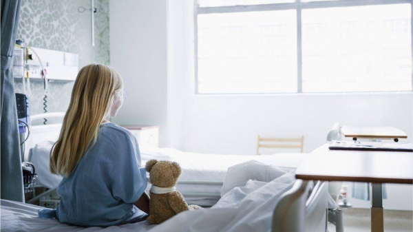 Young girl sitting on a hospital bed with a teddy bear next to her.