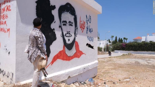 Samia stands by a mural of her son in their hometown of Kelibia. She said his friends, who regularly visit her, painted it. The drawing has his face, the date of his disappearance, a map of Tunisia and says 