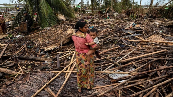 A Rohingya woman carries her baby next to her destroyed house at Basara refugee camp in Sittwe on May 16 after cyclone Mocha made landfall