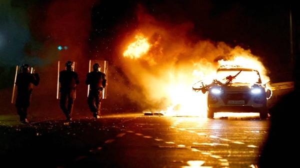 Riot police walk next to a burning police vehicle, near the scene of a suspected stabbing that left a few children injured in Dublin, Ireland