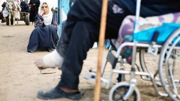 A woman sits on a dirt road, her chin resting on her hand. Out of focus in front of her is a man with an injured foot sitting in a wheelchair. — courtesy Reuters