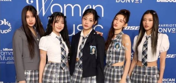 The five members of NewJeans stand against a blue and turquoise backdrop, dressed in variations of tartan school skirt with blazers and ties