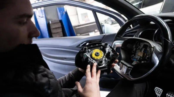 A man sat in a car's driving seat takes apart a steering wheel to replace a faulty airbag system
