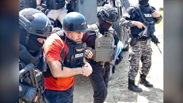 Ecuadorean drug lord 'Fito' wears a hard hat and an orange t-shirt underneath a bullet-proof vest. He is escorted by several people in black and camouflage clothes, also wearing bullet-proof vests