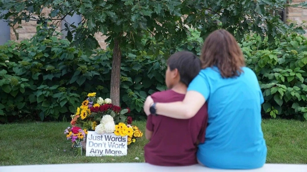 People sit across a memorial near boarded windows damaged in Wednesday's school shooting at Annunciation Catholic Church, Thursday, Aug. 28, 2025, in Minneapolis