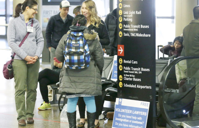 Asti Gallina, right, and Emily McDaniel, left, both volunteer law students, talk to a traveler as they staff a station near where passengers arrive on international flights at Seattle-Tacoma International Airport in Seattle on Tuesday. — AP